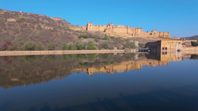 A wide shot of the historic Amer Fort in Jaipur, India, perfectly reflected in the calm waters of Maota Lake under a clear blue sky.