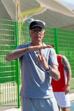 Senior referee in visor and glasses signals timeout with hands on outdoor basketball court during sunny game, focused and authoritative