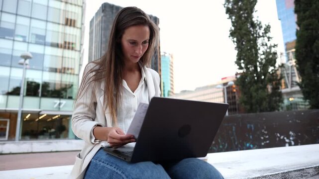 Young professional woman happily reviewing business documents and working on her laptop in a modern urban setting, symbolizing career success, remote work, and contemporary business