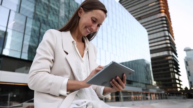 Young businesswoman working remotely outdoors, concentrating while using a digital tablet against the backdrop of a modern glass office building, symbolizing flexible work and connectivity