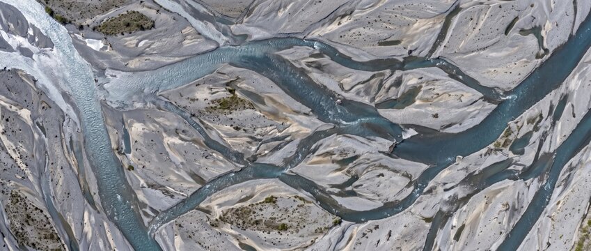 Aerial view of a braided river system with winding blue water channels, grey gravel bars, and sediment deposits forming an intricate, branching network across a wide, dry riverbed landscape