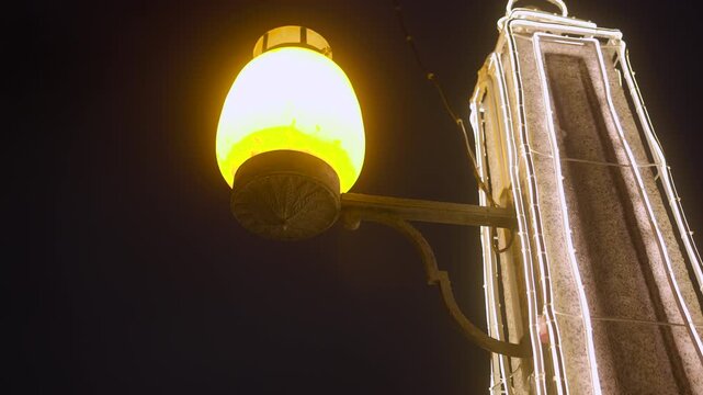 Glowing streetlamp beside illuminated stone tower at night with warm amber light, ornate metal bracket, bright white outline lighting, dark sky, and dramatic urban atmosphere