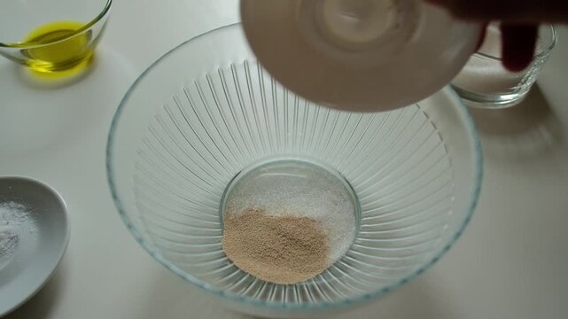Baker adding water sugar and yeast into glass bowl on kitchen table