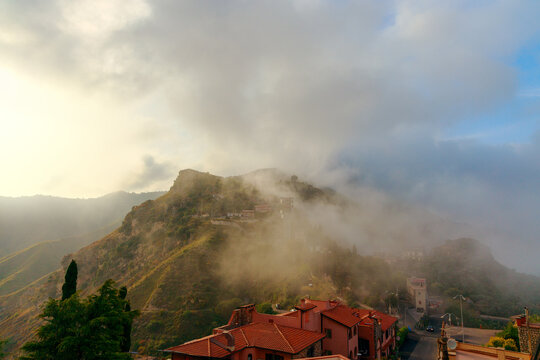 Golden light illuminates the rugged hillside landscape where traditional houses with terracotta roofs nestle amidst the rolling misty terrain