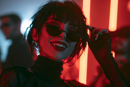Modern Vampire Woman in Gothic Mesh Costume and Sunglasses Posing at a Red Neon Halloween Nightclub