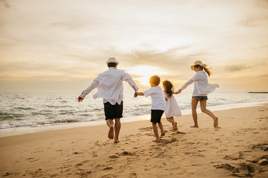 Back of happy Asian parents with their daughters enjoying playful at beach. Little girls bond with their mother and father running jumping and pretending to fly with arms wide open. Family fun in sun.