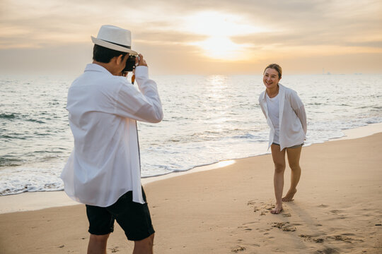 A couple in white attire celebrates love at sunrise by sea with boyfriend skillfully taking photos of his girlfriend. beach becomes canvas for their romance creating beautiful memories together.