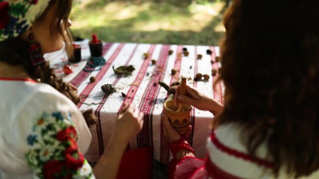 Ukrainian women perform witch rituals while dressed in vyshyvankas. They gather herbs and create patterns on a table in a natural setting. The focus is on their cultural practices.