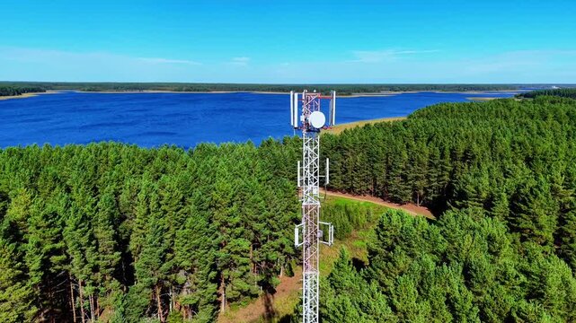 Aerial flight of telecommunications tower surrounded by dense green forest and blue lake