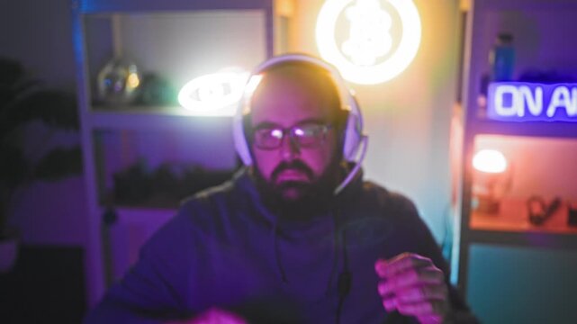 Bald hispanic man with beard and glasses wearing a headset and hoodie, adjusting a microphone with hand raised beside a neon on air sign in studio; concentration.