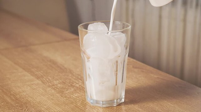 Pouring fresh plant based dairy free milk over ice cubes in tall clear glass on wooden table. Preparation of iced coffee or iced matcha latte drink.
