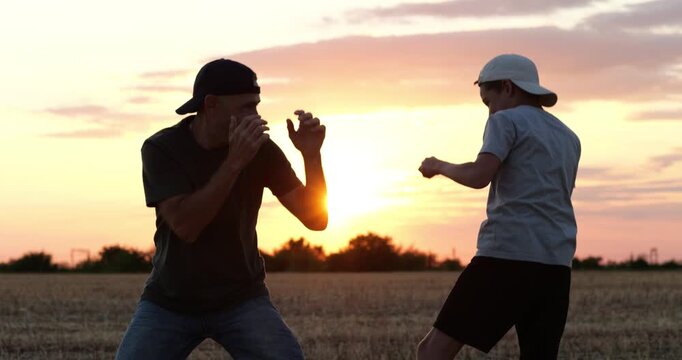 Father and son playfully spar in wheat field at sunset, smiling and enjoying time together. Fun boxing game creates warm family moment about bonding, laughter, connection and shared outdoor leisure