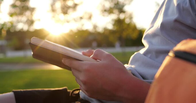 Child enjoys reading book outdoors in peaceful park at sunset. Hands hold open notebook beside orange backpack, capturing quiet study, learning, and relaxation. Warm golden light creates calm