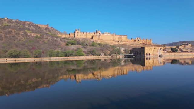 A wide shot of the historic Amber Fort in Jaipur, India, showing its majestic sandstone walls reflected in the calm waters of Maota Lake under a clear blue sky.
