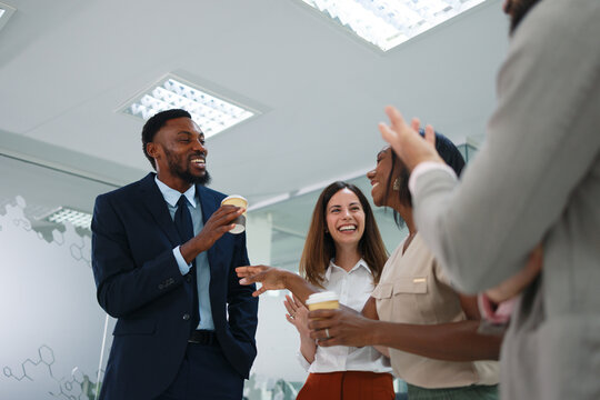 Diverse business people smiling and laughing while holding coffee cups during a break in a bright modern office, enjoying informal conversation and building camaraderie