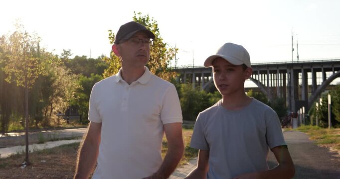 Father and son walk and talk in urban park at sunset. They converse near bridge in warm evening sunlight during golden hour. Family connection supports parenting, communication, and togetherness.