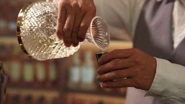 Close-up of a bartender pouring clear spirits from a cut glass decanter into a metal jigger at a warm, elegant bar.