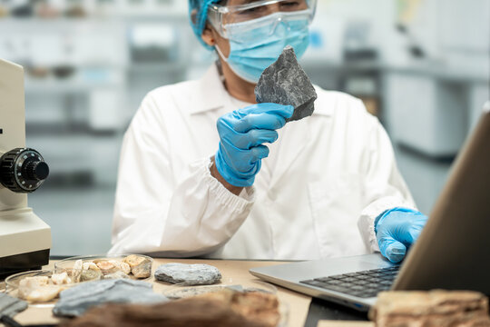 Geologist in protective gloves and mask examines a rock sample beside fossils, a microscope and laptop in a modern laboratory.