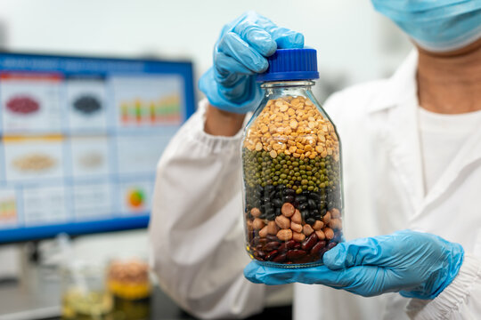 Food scientist in protective gloves holds a glass jar of mixed legumes in a laboratory, with a computer screen showing test results in the background.