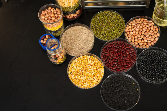 Top view of assorted dried beans, lentils, peas and grains in clear bowls and jars on a black background with empty copy space.