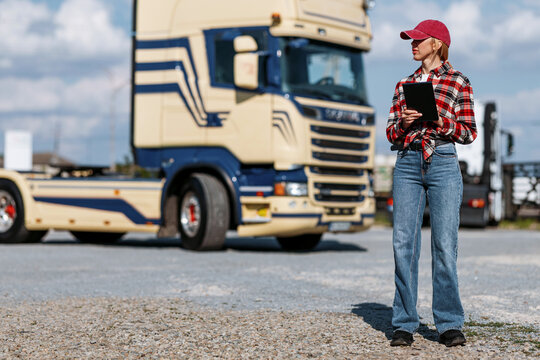 A female truck driver in casual attire stands with a tablet, overseeing operations next to a large commercial vehicle