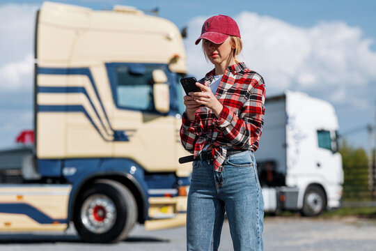 A female trucker in a plaid shirt and baseball cap uses her smartphone outdoors near two large semi-trailer trucks on a sunny day