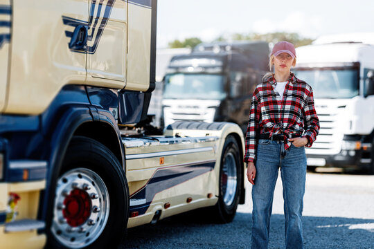 A young woman wearing a plaid shirt and denim jeans stands confidently next to a large semi-trailer truck in a parking lot