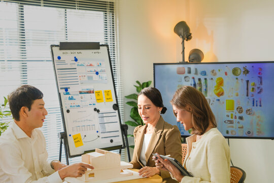 The man leans back on his chair while the women continue the discussion, in a daylit creative studio, an Asian architecture and design team collaborating on an environmental project.