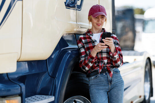 A female trucker in casual attire checks her phone for messages or navigation while taking a break next to her large commercial vehicle
