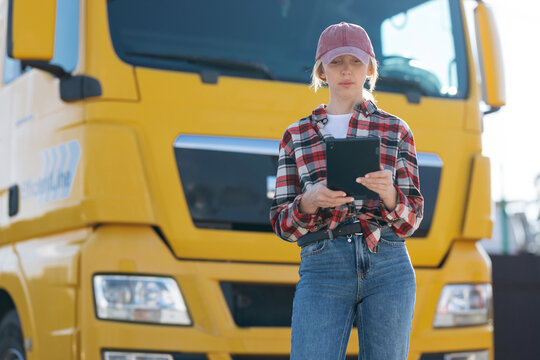 A woman wearing a plaid shirt and baseball cap stands in front of a large commercial vehicle while reviewing shipment details on a handheld device