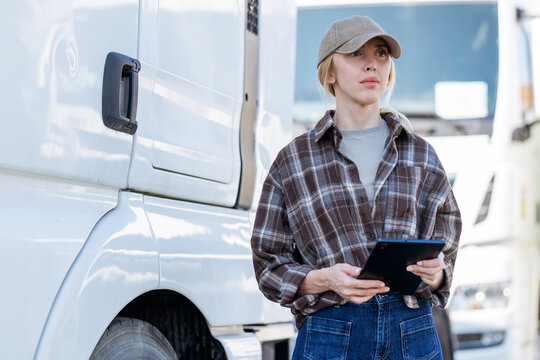 The professional woman wears a plaid shirt and baseball cap as she reviews logistics data on a handheld screen during a break at the shipping yard