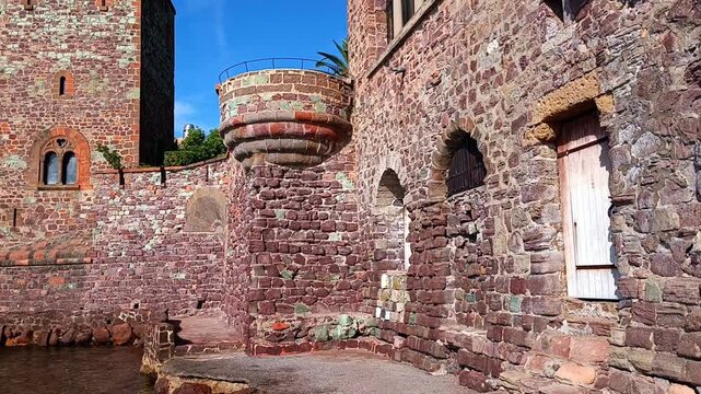 A tall red brick tower with small arched windows and a circular turret stands attached to a stone wall under a clear blue sky at a historic French castle at Chateau de La Napoule in Mandelieu-la-Napou