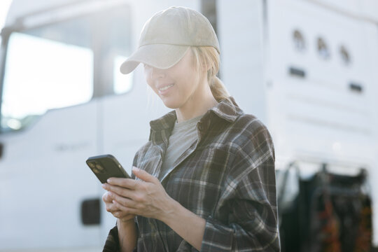 A smiling person checks a mobile device while standing in front of a large white vehicle during a bright day with soft natural lighting