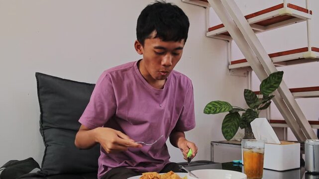 Young man eating batagor with fork and knife while sitting at living room table