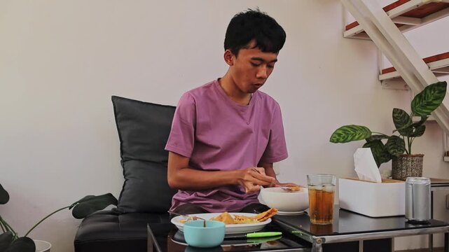 Young Asian man eating batagor kuah with spoon and fork at home