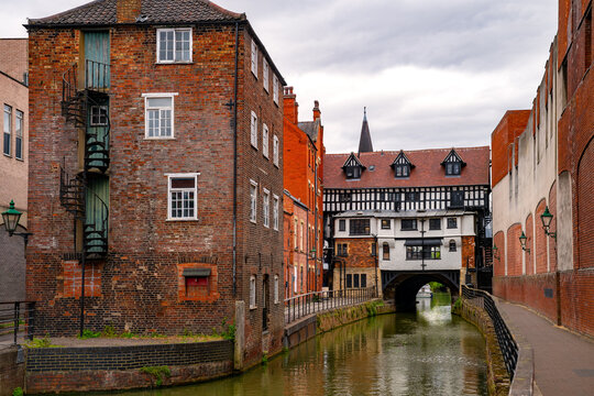 Historic High Bridge spanning the River Witham in Lincoln, England, framed by old brick buildings and waterside architecture beneath an overcast summer sky.