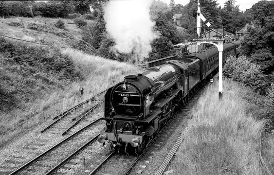 Historic steam locomotive leaving Goathland Station in North Yorkshire, England, beneath a traditional semaphore signal. Black and white summer railway scene.