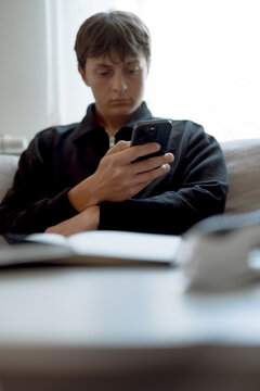 Young man sits indoors, focused on smartphone. Soft light filters through window behind him. Dark jacket contrasts with muted background tones. Expression shows concentration or mild distraction