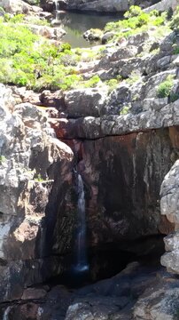 Narrow waterfall cascading over layered granite cliff into shaded plunge pool inside steep sunlit gorge, water splitting into trickles and spraying mist below rock ledges