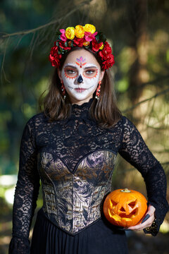 Halloween beauty portrait of woman in forest with Halloween pumpkin in hand. Santa Muerte makeup.