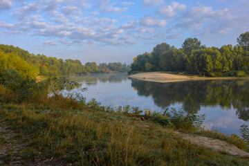 A scenic, peaceful river flows gently under a partly cloudy blue sky, its calm surface perfectly reflecting the lush green forests and sandy banks lining both sides during a bright morning. © Александр Арендарь