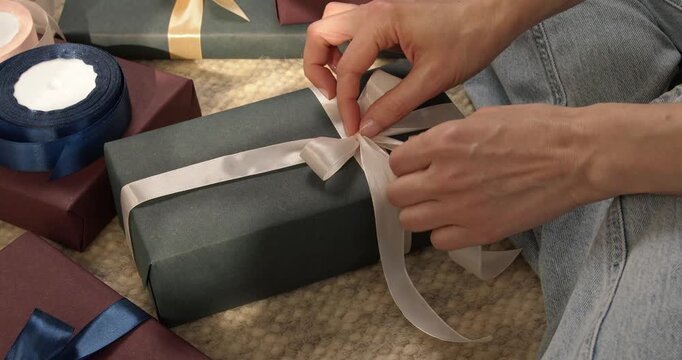 Woman hands tying white satin ribbon on gift box with other presents around close up video.