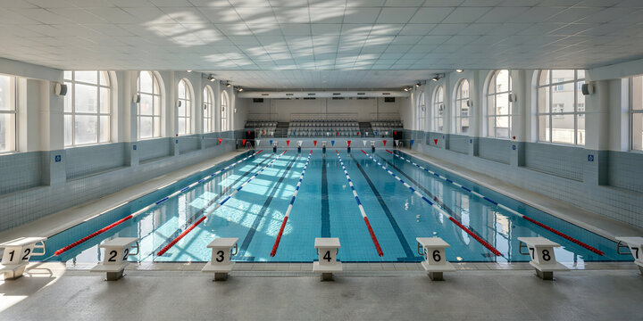 Indoor swimming pool with blue water, lane markers, numbered starting blocks, and arched windows in a bright, spacious aquatic facility hall