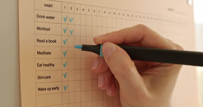 Close up of woman hand ticking various tasks with blue marker on paper habit tracker.