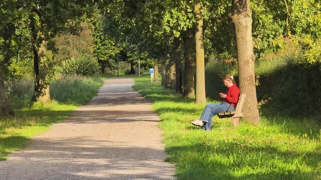 Woman in a red sweater sits on a bench in a park during a sunny day