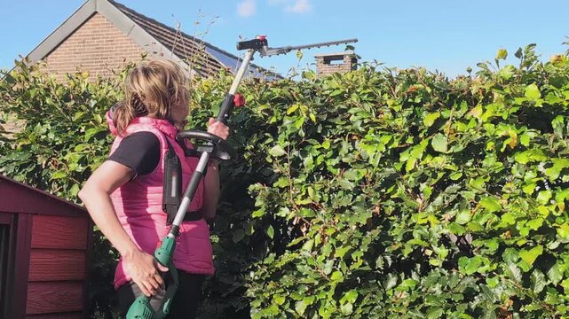 Woman trims bushes with electric trimmer in backyard during sunny day