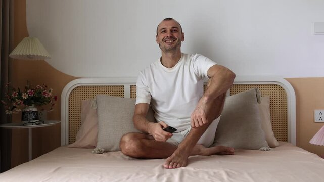 Young man sitting on bed, holds remote control, watching TV and smiling.