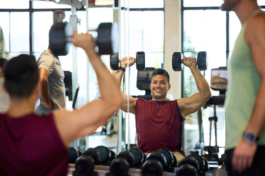 Man lifting dumbbells in gym mirror. He raises weights overhead with focused strength. Smiles with satisfaction and determination. Shoulder Press. Office in-house gym