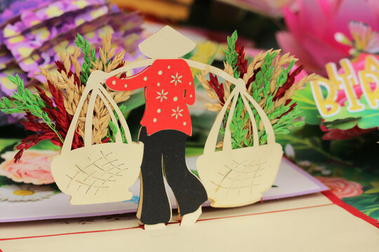Intricate 3D popup paper card depicting a traditional Vietnamese woman wearing a conical hat and carrying baskets with rice crops in Hoi An, Vietnam.