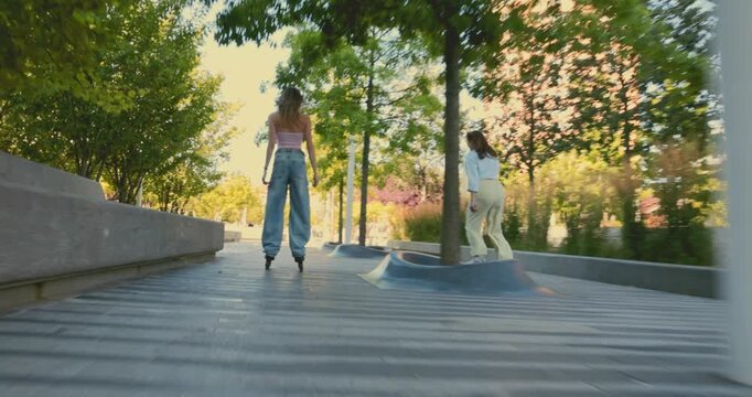 Two young women rollerblade through leafy urban park as low follow camera captures playful shared motion. Friendship, active lifestyle and urban recreation concept.